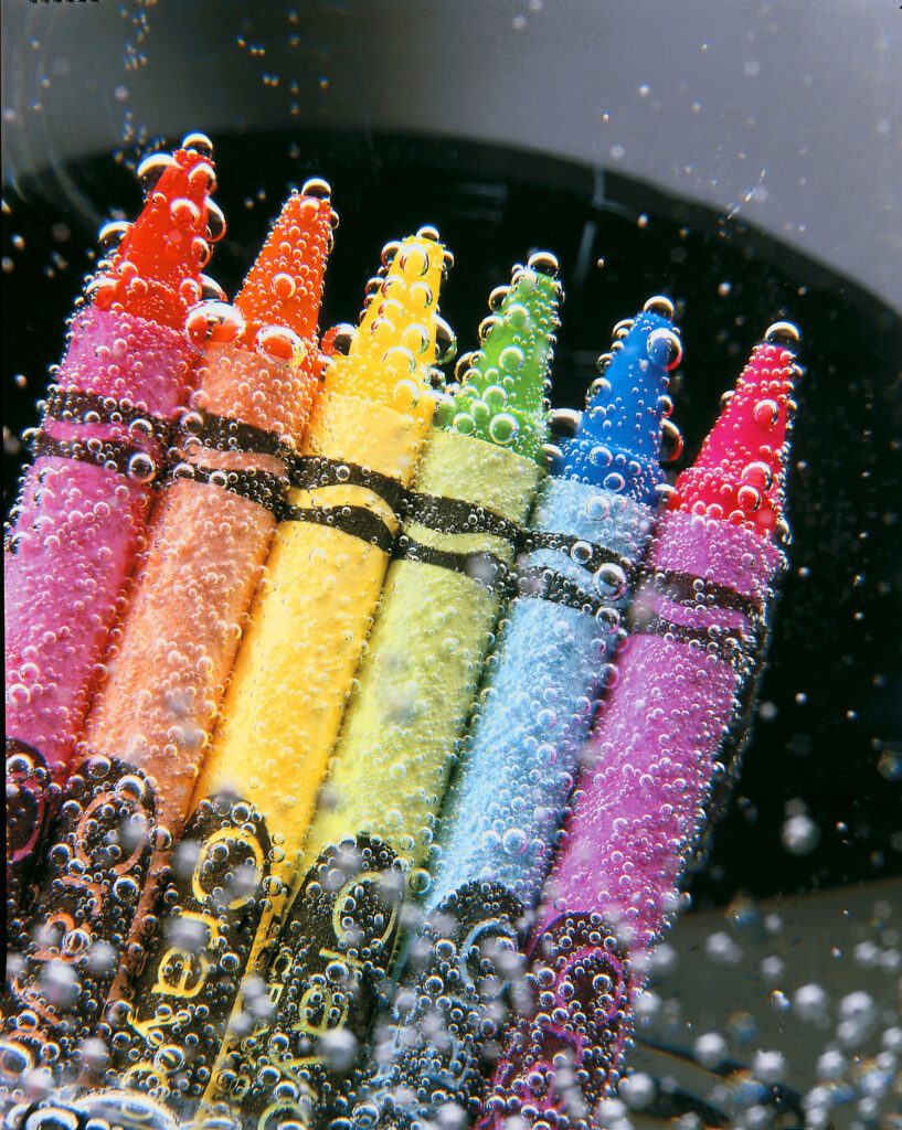 Rainbow of wax crayons covered in water bubbles against a dark background