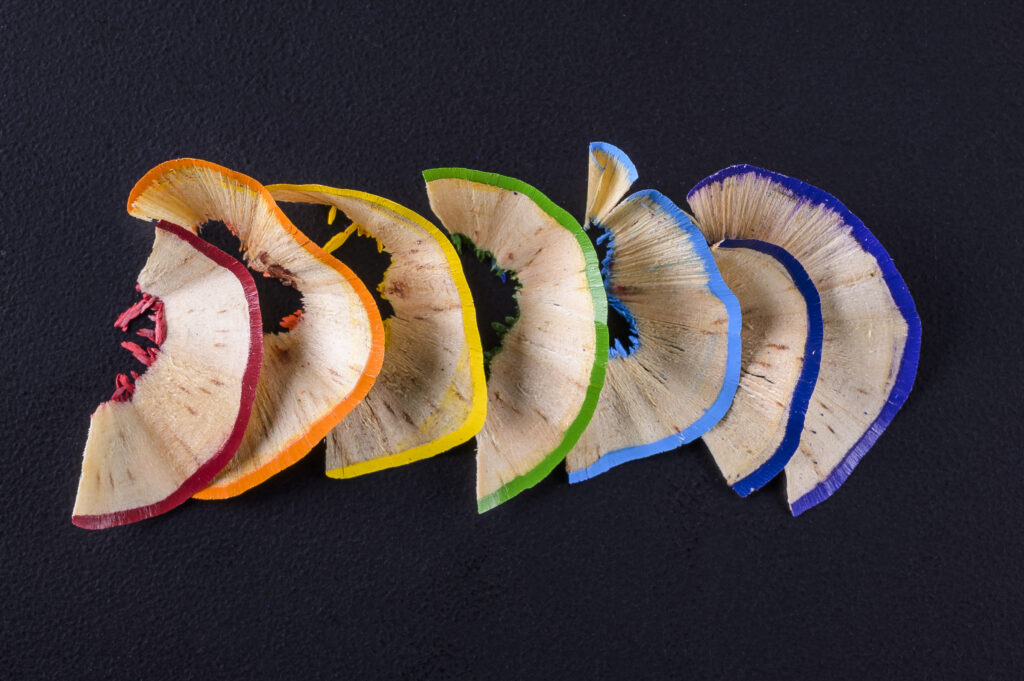 Colored pencil shavings curled in a rainbow arc, from red to blue, on a dark background