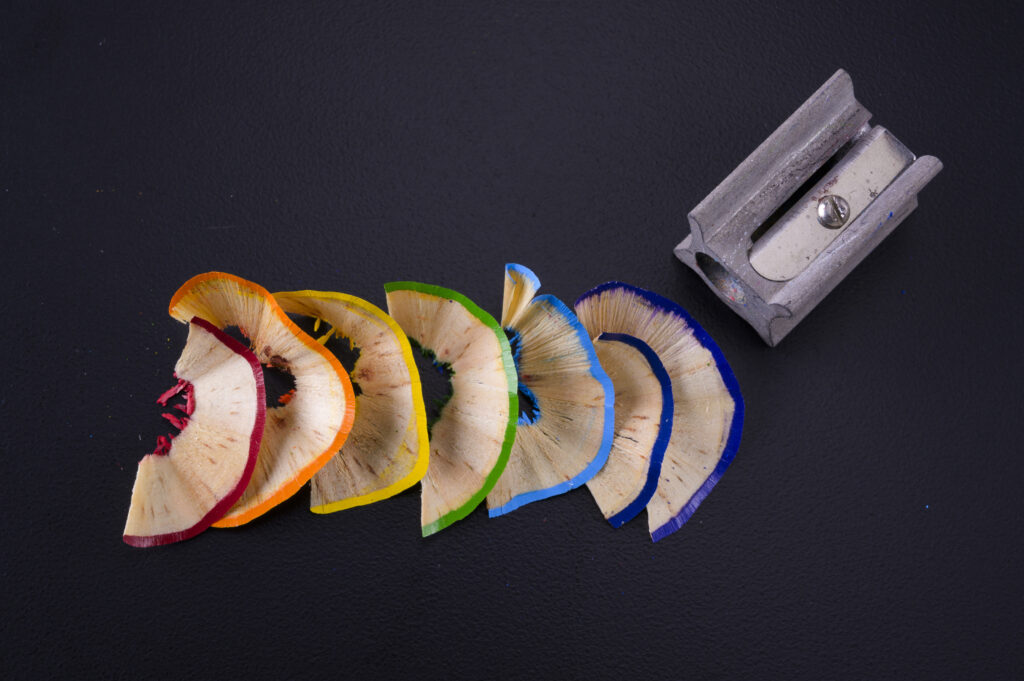Rainbow of colored pencil shavings beside a gray pencil sharpener on a dark background