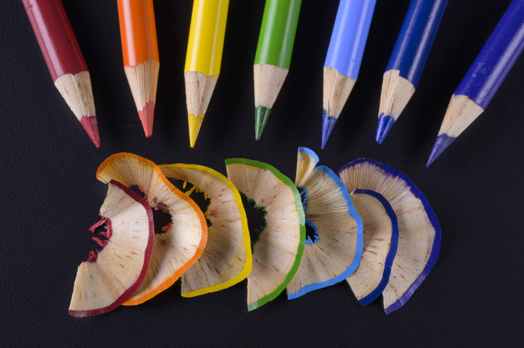 Seven colored pencils above a matching rainbow arc of their shavings on a dark background