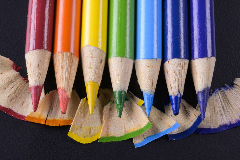 Close-up of the sharpened tips of seven colored pencils, red through blue, above their shavings